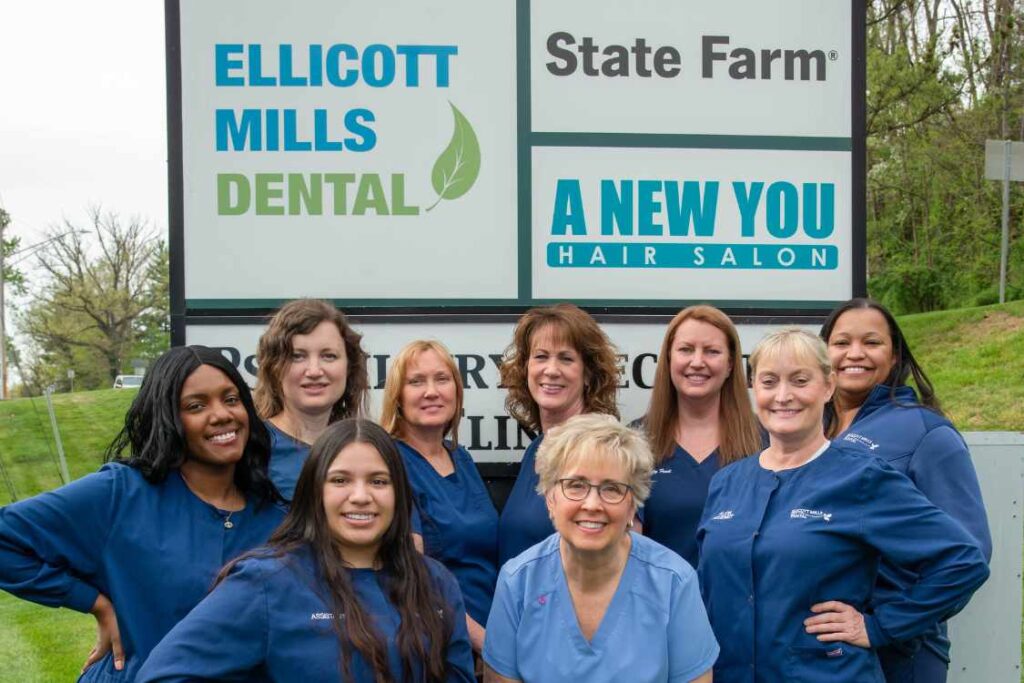 9 women dressed in blue scrubs posing in front of the Ellicott Mills Dental business sign.