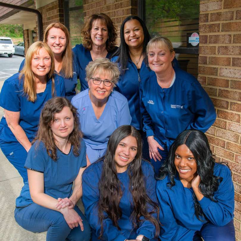 9 female Ellicott Mills Dental staff members posing in blue scrubs outside of the office next to a brick wall.